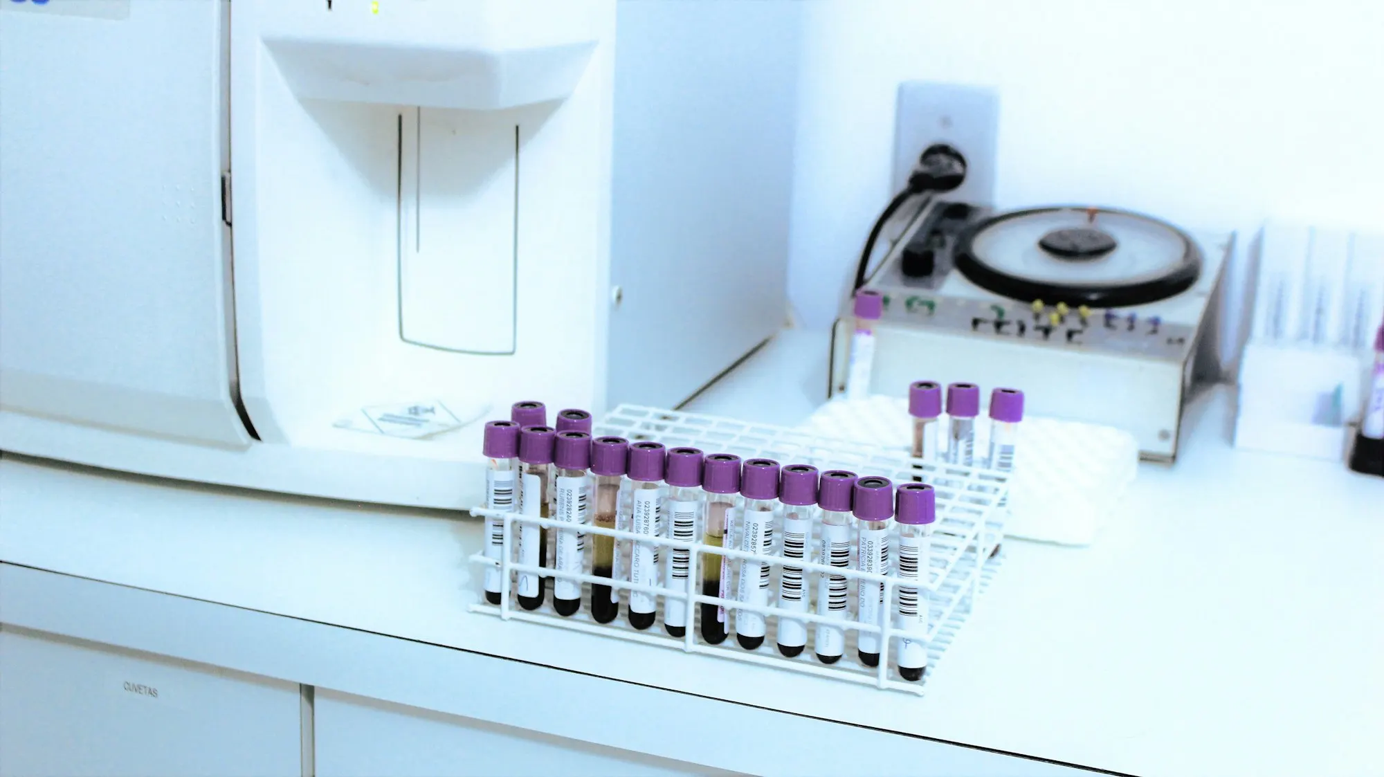 Portoir de tubes de prélèvement à bouchon violet posé près d'une centrifugeuse sur un plan de travail de laboratoire blanc.