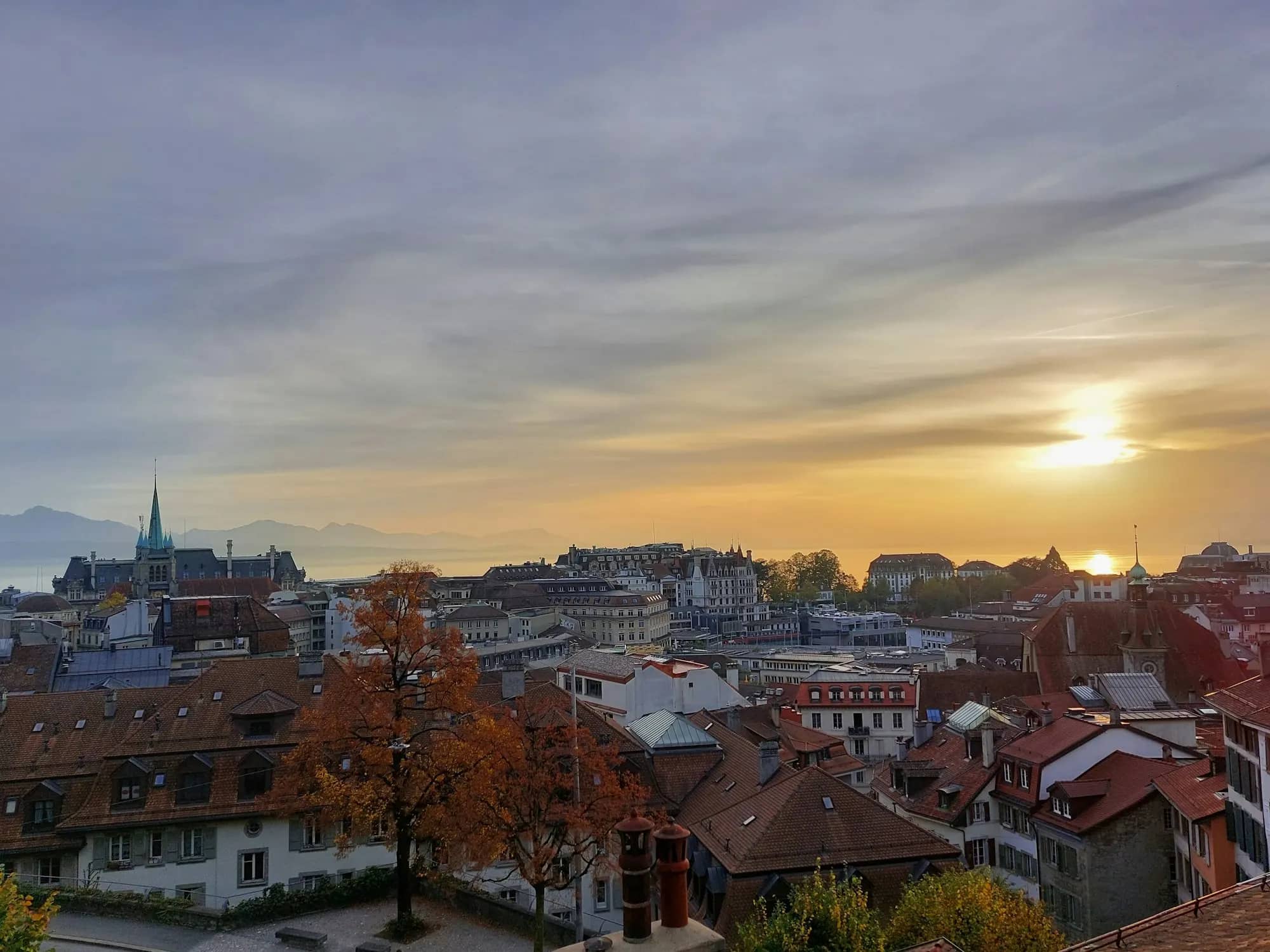 Vue sur les toits de Lausanne au coucher du soleil, avec la cathédrale à gauche et le Léman à l'horizon.
