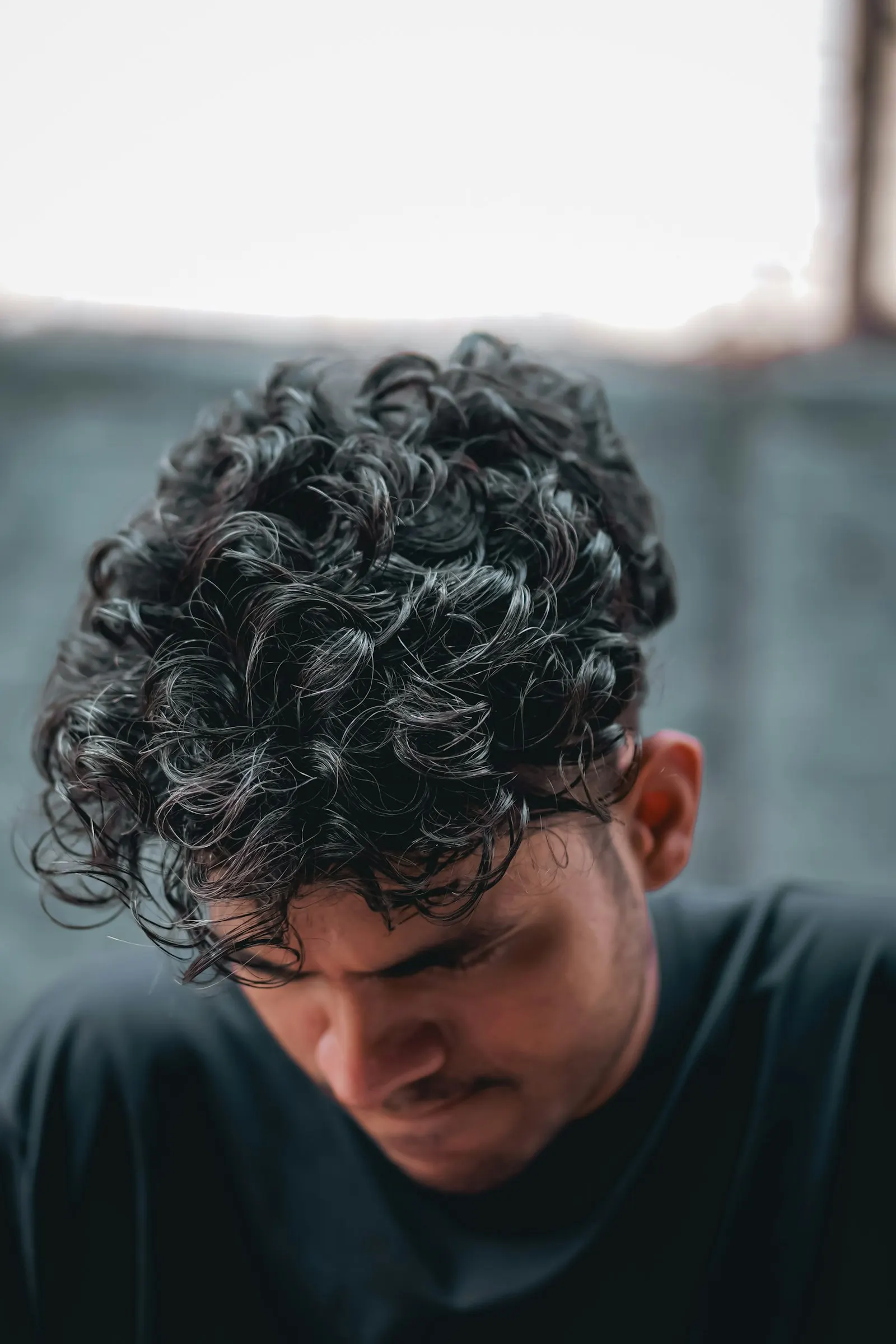 Homme tête baissée, vu de face légèrement en plongée : le sommet du crâne et ses cheveux bouclés occupent le haut de l'image.